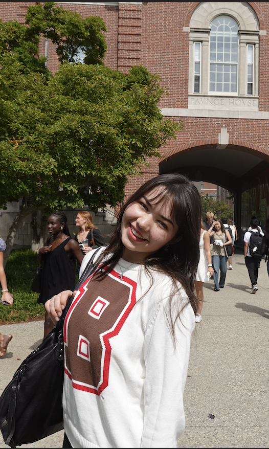 Isabel standing with her back to the Faunce Arch, wearing a white sweatshirt with the Brown logo on it as well as a big smile!