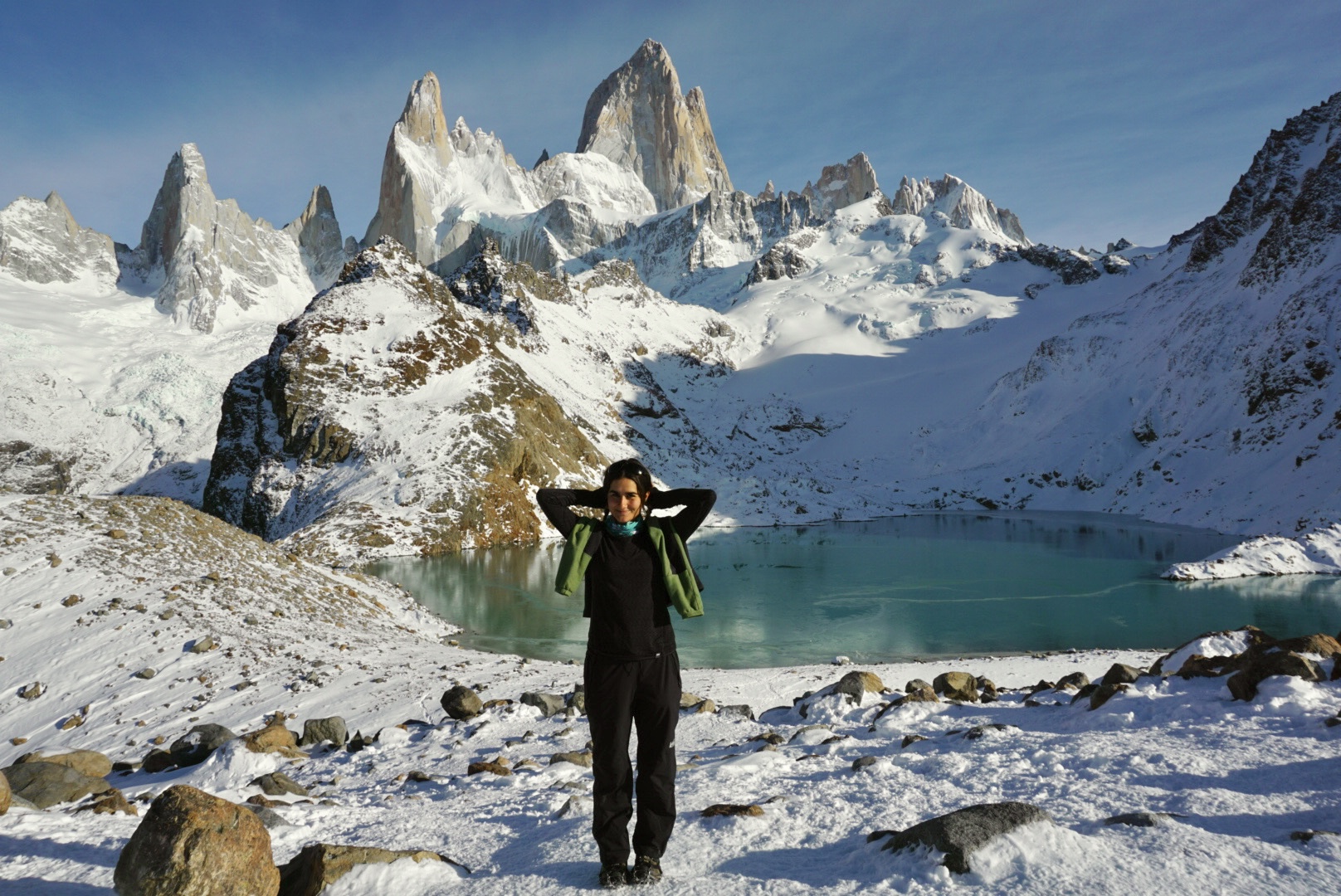 Isabel standing in front of a glacial lake, with her hands behind her head, looking accomplished. There are mountains in the background rising up over her.